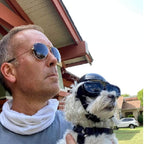 A man wearing round aviator sunglasses and a white neck gaiter holds a small white dog wearing a black aviator helmet and goggles outdoors in front of a house
