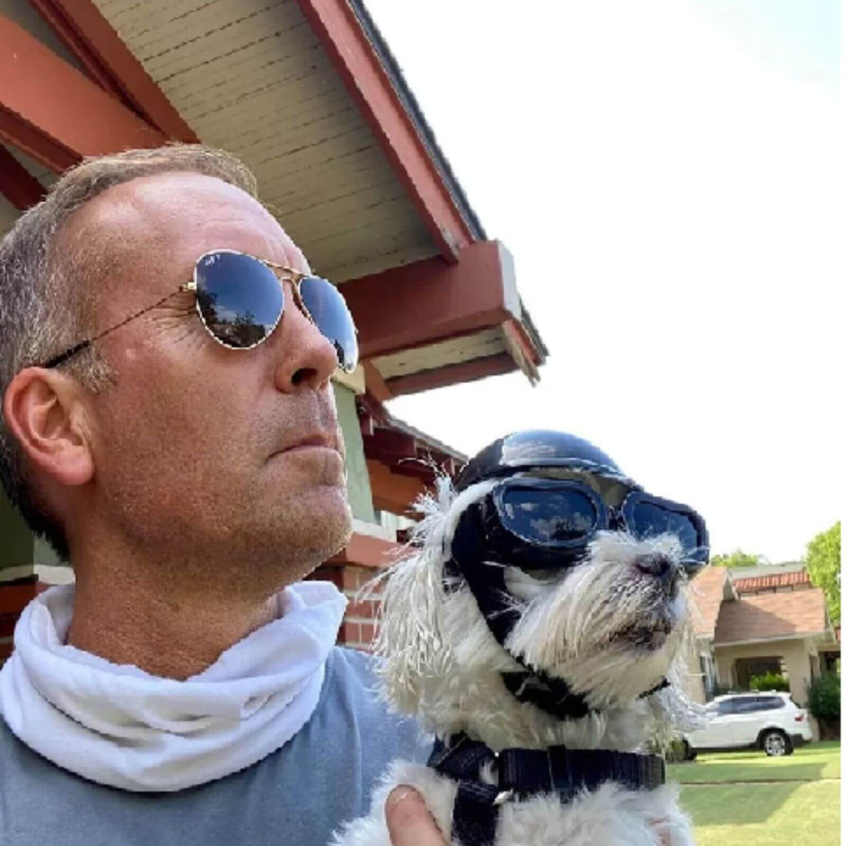 A man wearing round aviator sunglasses and a white neck gaiter holds a small white dog wearing a black aviator helmet and goggles outdoors in front of a house