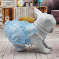 A white dog-shaped decorative sculpture or costume display wearing a fluffy light blue petal dress and a white harness, shown indoors on a wood floor with shelves and decor in the background
