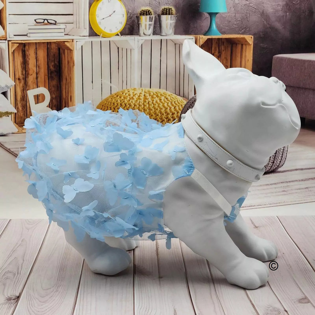 A white dog-shaped decorative sculpture or costume display wearing a fluffy light blue petal dress and a white harness, shown indoors on a wood floor with shelves and decor in the background