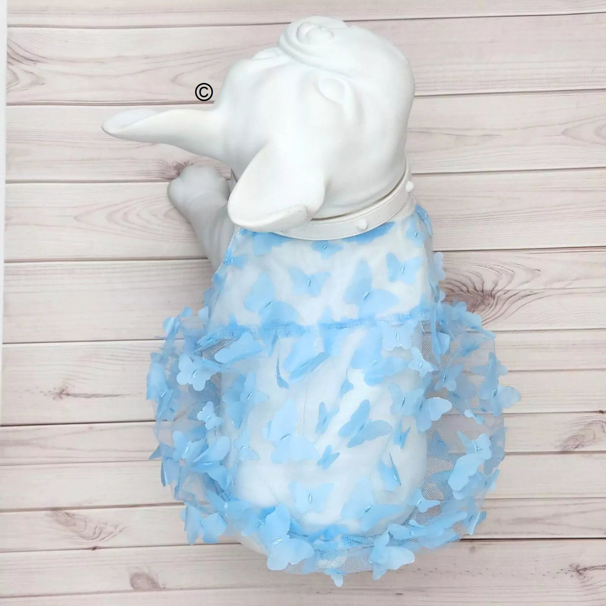 A white dog statue or figurine wearing a fluffy light blue tulle dress covered with small blue butterfly embellishments, placed on a light wood plank background.