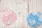 Three ruffled pet harnesses in pink, white, and blue, displayed on a light wood plank background