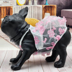 A black dog statue wearing a sheer white pet dress with pink floral appliqués and pink polka-dot tulle trim, posed on a light wood floor in a styled indoor room setting