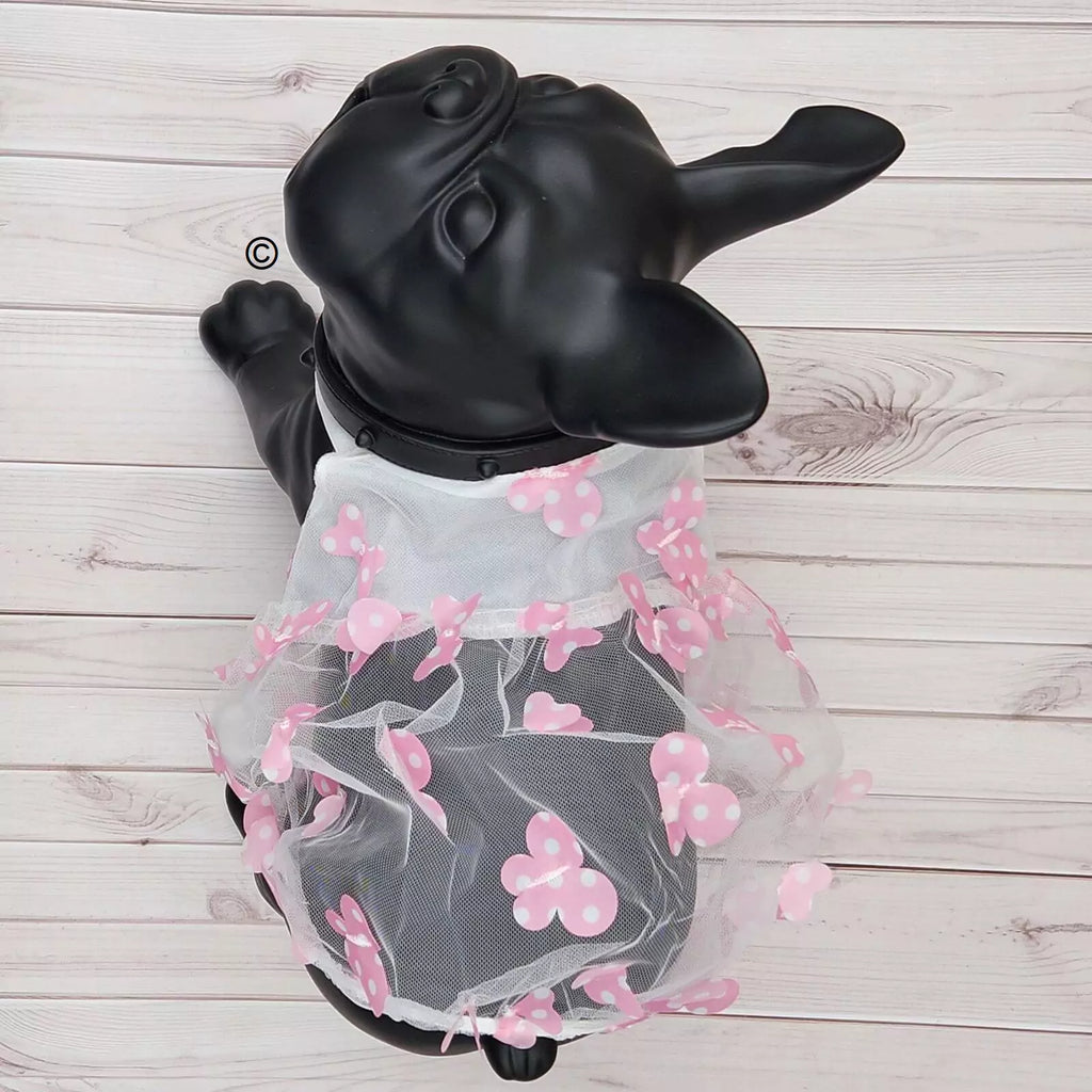 A black French bulldog dog coat dress displayed on a black dog mannequin, featuring a sheer white tulle skirt decorated with pink cherry blossom flowers, set against a light wood plank background.