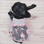 A black French bulldog dog coat dress displayed on a black dog mannequin, featuring a sheer white tulle skirt decorated with pink cherry blossom flowers, set against a light wood plank background.