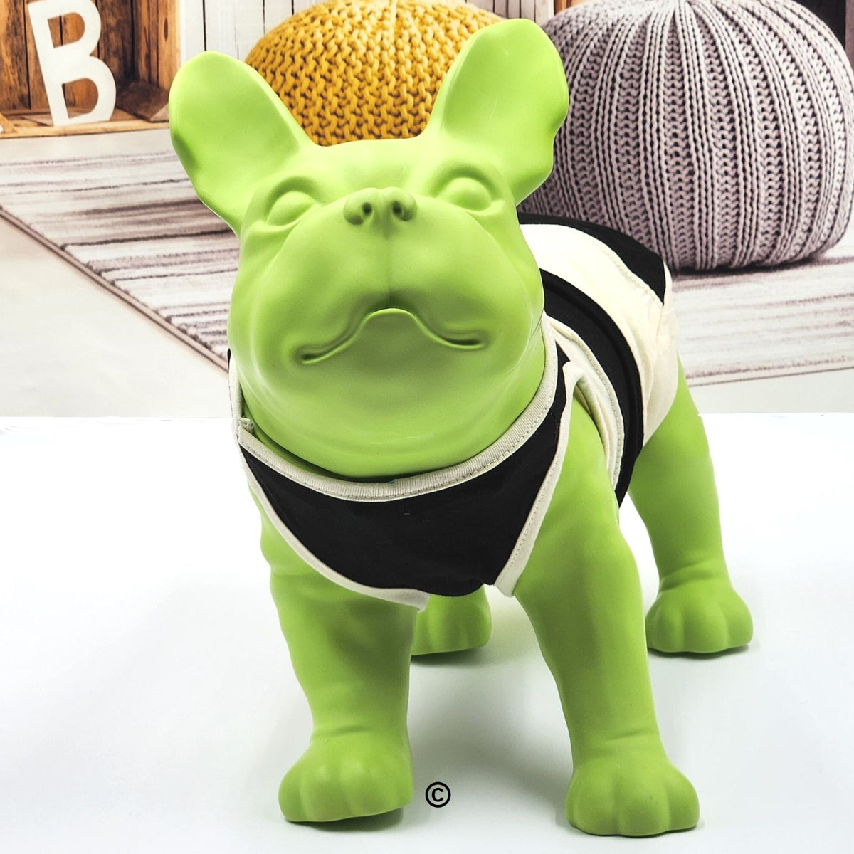 A bright green French Bulldog toy wearing a black and white harness, standing on a white surface with a cozy, styled background featuring a yellow knit pumpkin and a gray knit pillow.