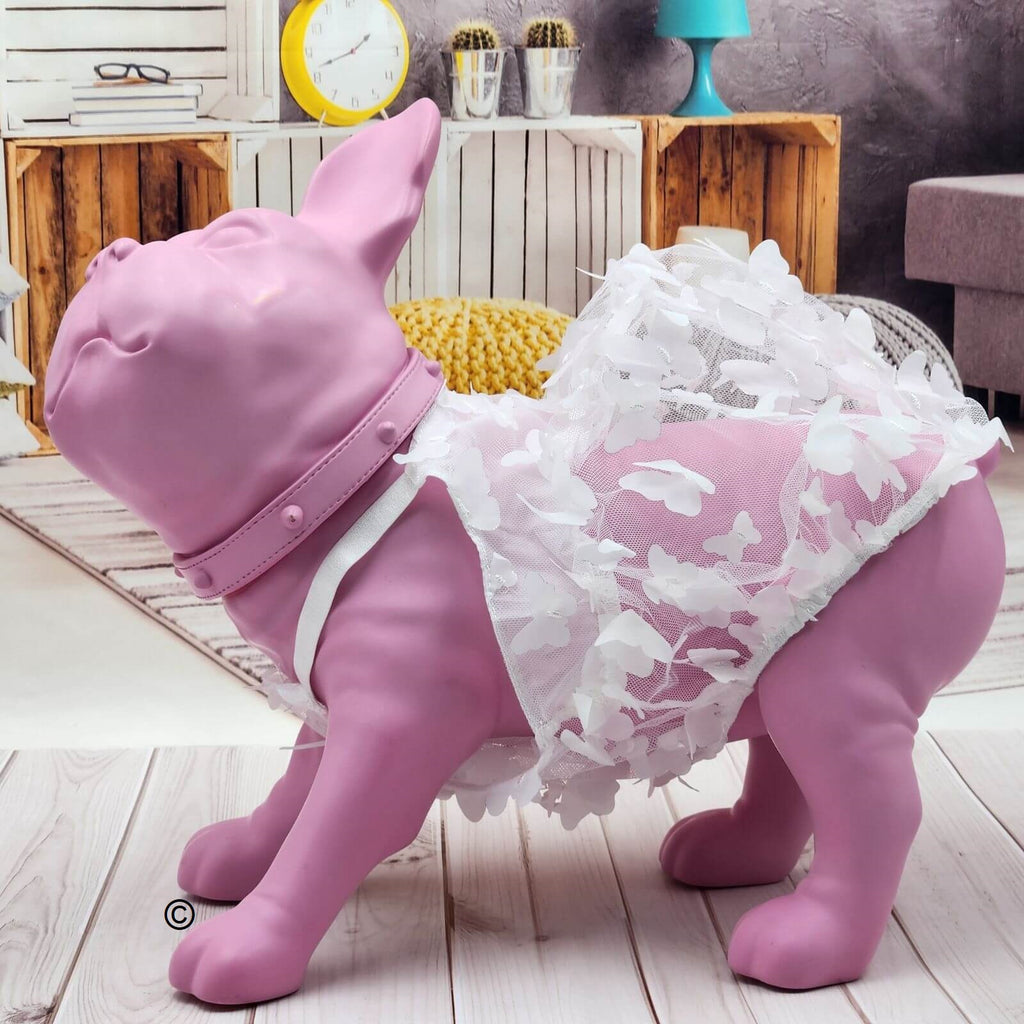 A pink dog-shaped decorative statue wearing a white floral lace dress and collar, posed on a light wood floor in a styled room setting.