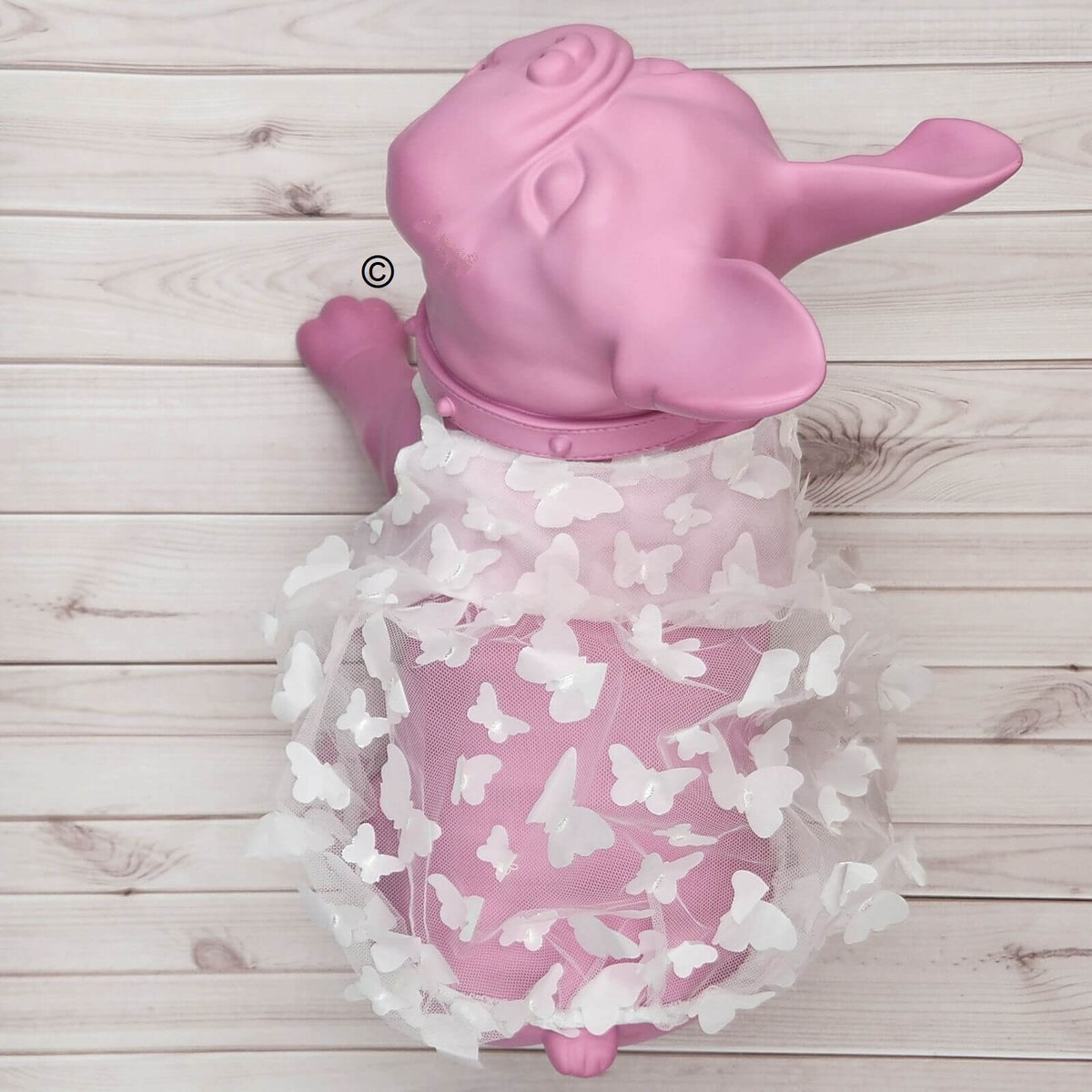 Pink elephant-shaped wall art or decorative figure with a white sheer tulle skirt covered in white butterfly appliqués, shown against a light wood plank background.